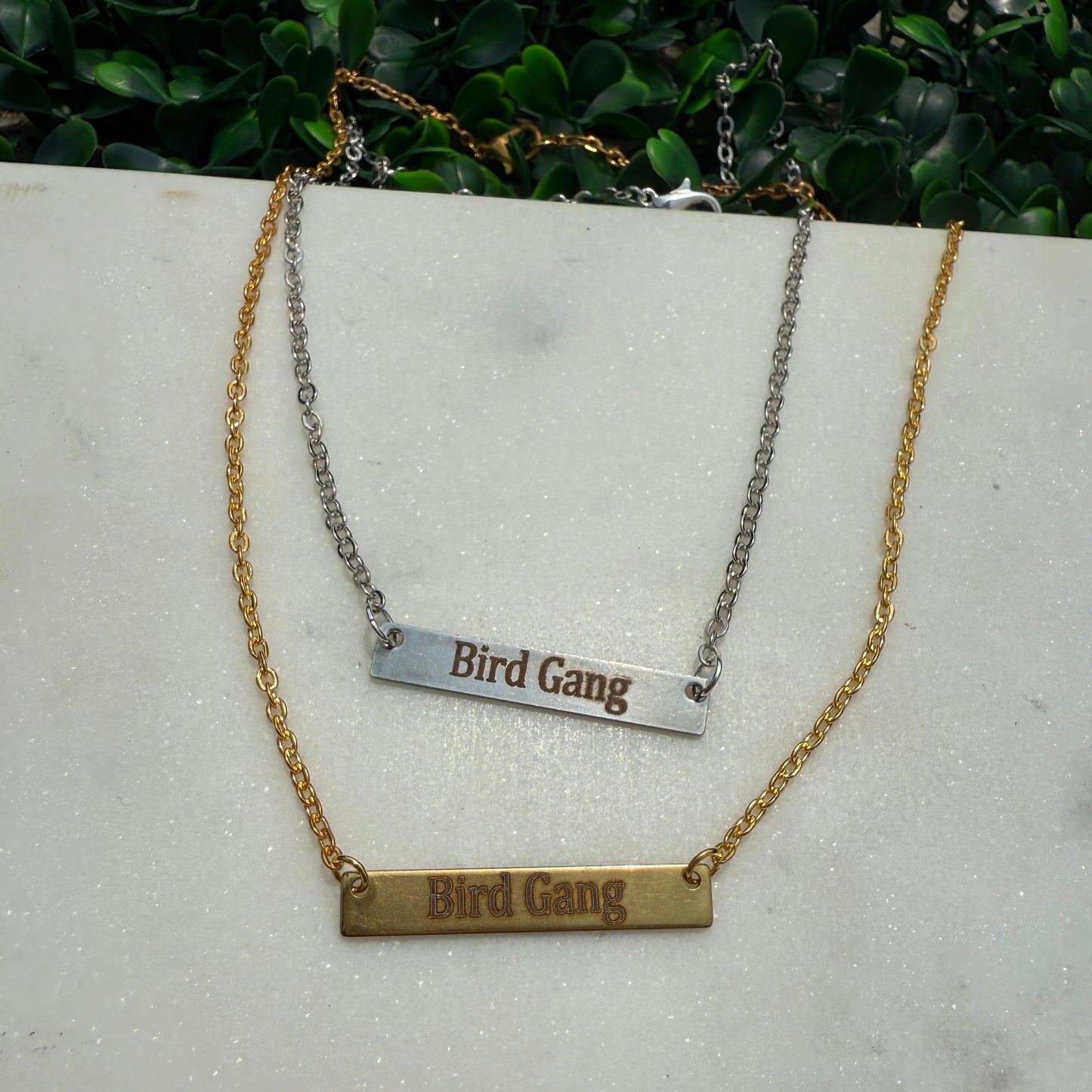 Two necklaces with 'Bird Gang' engraved on them, one gold and one silver, on a light gray surface.
