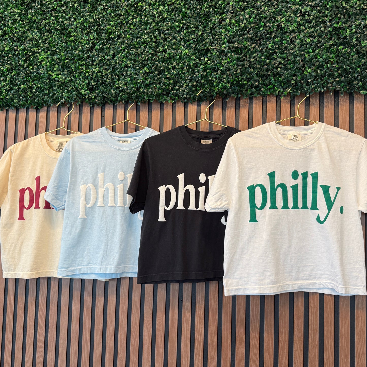 Four t-shirts with 'philly' text in different colors hanging against a green leafy background.