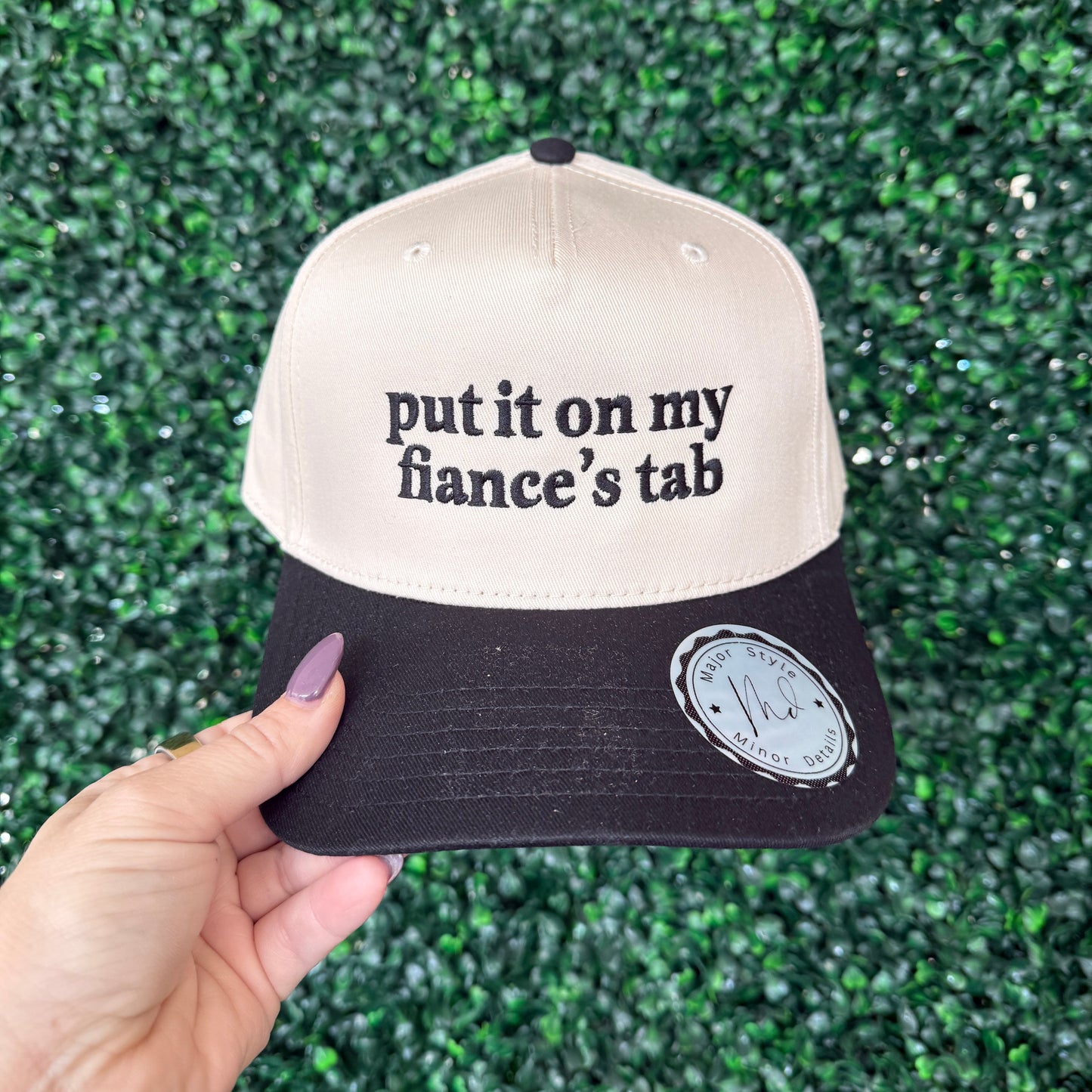 Cap with text 'put it on my fiancé's tab' held against a green textured background