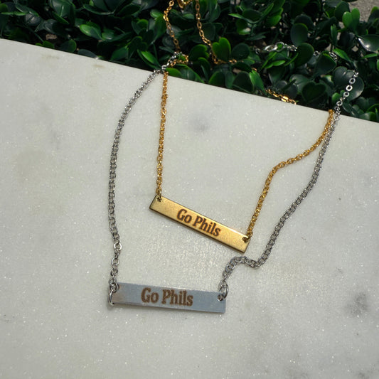 Two gold and silver bar necklaces with 'Go Phillies' text on a concrete surface.