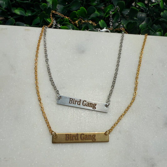 Two necklaces with 'Bird Gang' engraved on them, one gold and one silver, on a light gray surface.