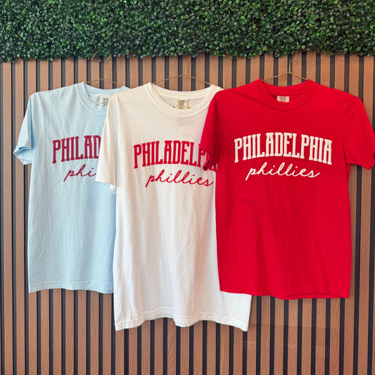 Red, white, and light blue t-shirts with 'Philadelphia Phillies' text on a wooden surface.