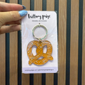 Gold pretzel-shaped keychain on a card with 'brittany paige' branding against a striped background.