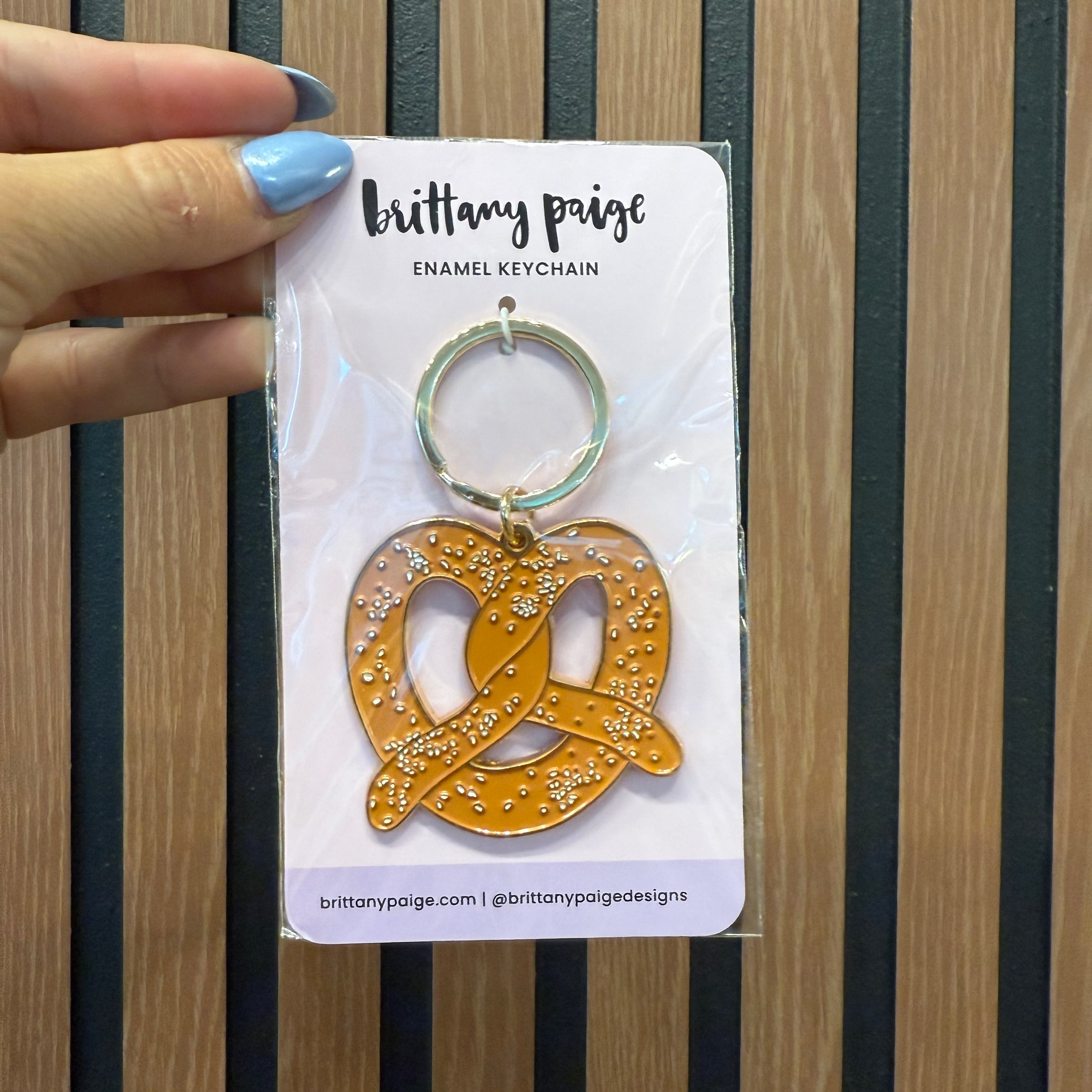 Gold pretzel-shaped keychain on a card with 'brittany paige' branding against a striped background.