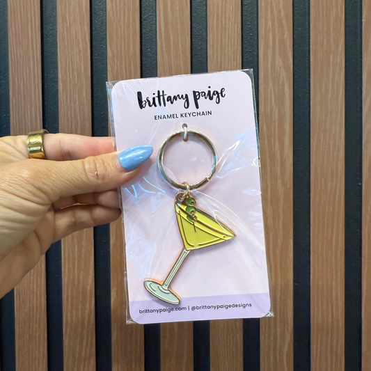 Yellow cocktail glass keychain on a clear card held by a hand with blue nail polish against a striped background.