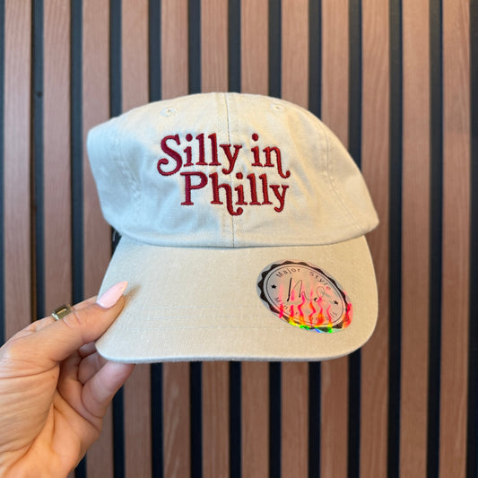 Beige cap with 'silly in Philly' text held against a wooden background