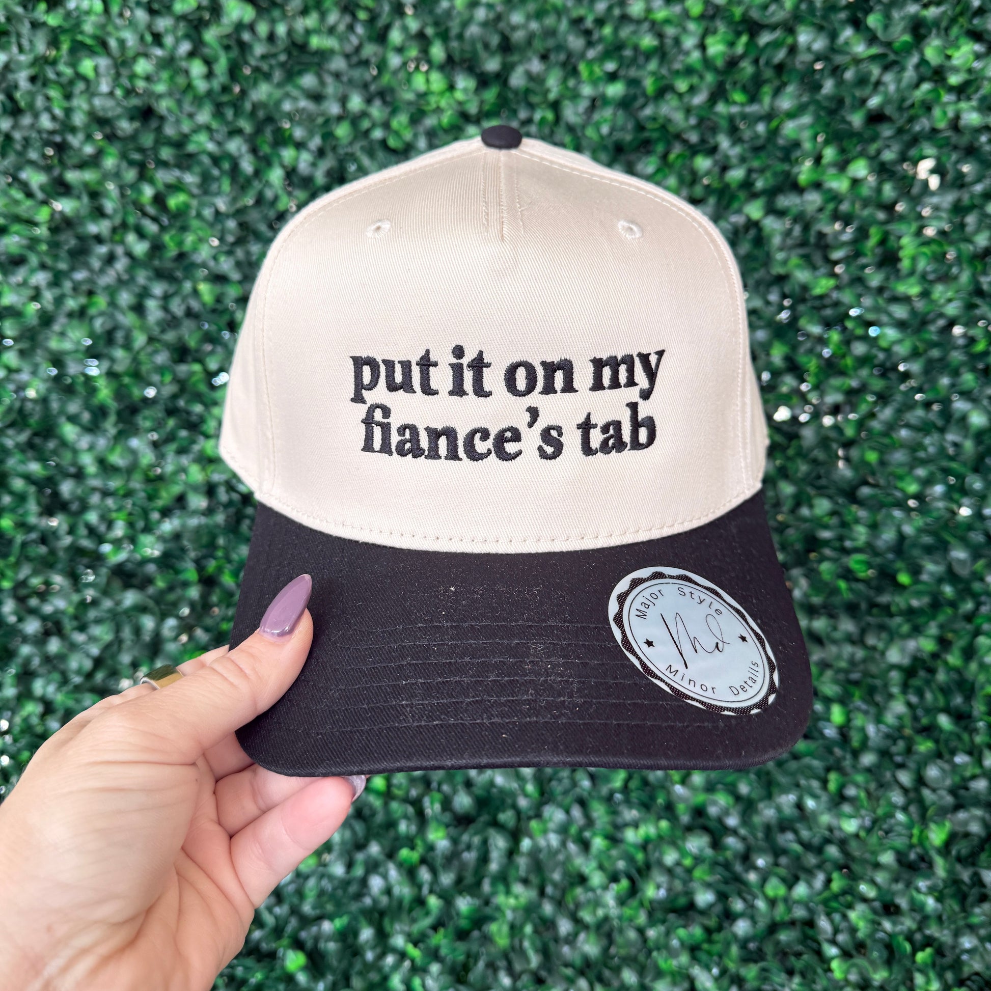 Cap with text 'put it on my fiancé's tab' held against a green textured background